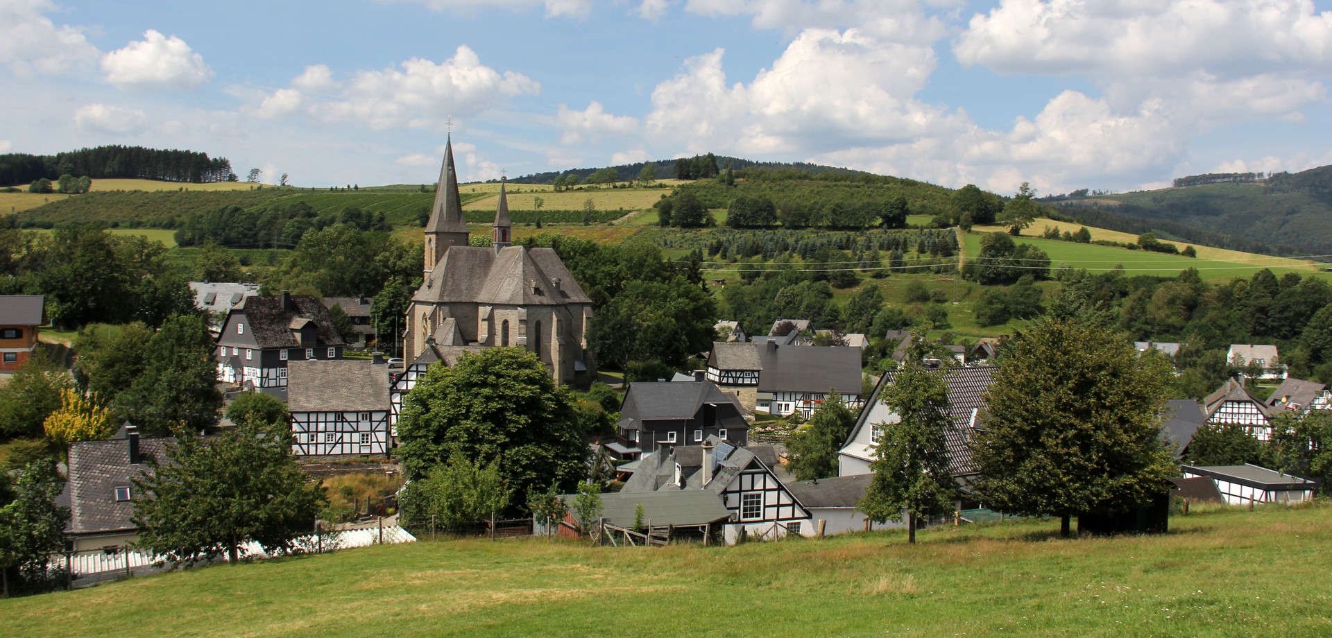 Dorf mit Kirche und Fachwerkgebäuden in einer hügeligen Landschaft